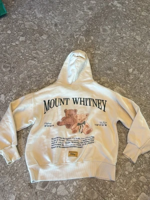 Beige hoodie med Mount Whitney-tryck - Beige hoodie med huva och känguruficka. Stor text 'Mount Whitney' på ryggen och två nallebjörnar tryckta under. På framsidan står det 'California' och en liten nalle. Mjuk bomullskänsla och avslappnad passform, perfekt för en chill look.