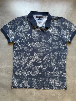 Blå mönstrad piké från Tommy Hilfiger - Säljer en blå pikétröja från Tommy Hilfiger i slim fit med vitt paisley-inspirerat mönster. Tröjan har klassisk krage, korta ärmar och knappar vid halsen. Perfekt för dig som gillar en unik och stilren look.
