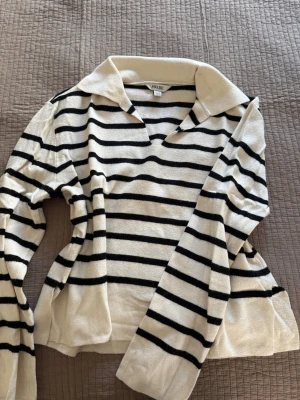 Randig stickad tröja med krage - Säljer en vit stickad tröja med svarta horisontella ränder från AWARE. Köp på Vero Moda. Tröjan har en bred krage och v-ringad hals, samt långa ärmar. Slits nere vid midjan. Perfekt för en avslappnad och trendig look.