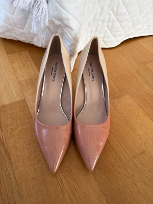Beige och rosa pumps med krokopräglad yta - Säljer ett par snygga pumps från ashton aubry i två olika nyanser av beige med glansig krokopräglad yta. Skorna har spetsig tå och smal klack på ca 9cm som ger en elegant look. Perfekta för dig som vill sticka ut med stilrena detaljer och trendig färgkombination.