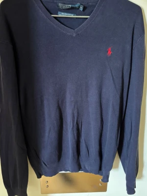 Mörkblå tröja med V-ringning Polo Ralph Lauren - Klassisk mörkblå tröja från Polo Ralph Lauren med V-ringning och röd broderad logga på bröstet. Tillverkad i mjukt material och har långa ärmar samt ribbade muddar vid ärmslut och nederkant. Perfekt för en stilren look. 