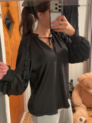 Svart blus med volangdetaljer - Snygg svart blus med långa puffärmar och volangdetaljer längs axlarna. Blusen har knytband vid halsen och smock vid ärmsluten för en trendig look. Perfekt att styla med både jeans och kjol.