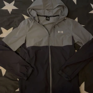 Grå och svart vindjacka från Under Armour - Snygg tvåfärgad vindjacka från Under Armour i grått och svart. Jackan har huva, dragkedja framtill och elastiska muddar vid ärmsluten. Perfekt för träning eller när du vill ha en sportig look. Under Armour-logga på bröstet.