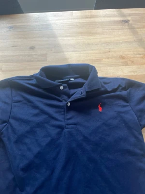 Marinblå pikétröja från Ralph Lauren - Klassisk marinblå pikétröja från Polo Ralph Lauren med röd broderad logga på bröstet. Tröjan har krage, knappar framtill och är tillverkad i mjuk bomull. Perfekt för en stilren och avslappnad look.