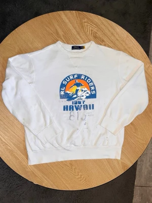 Polo Ralph Lauren RL Surf Riders Sweatshirt Vit – Storlek LG - Säljer en Polo Ralph Lauren sweatshirt i vit färg med klassiskt RL Surf Riders-tryck framtill.  Retroinspirerat tryck med Hawaii-motiv och RL-logga, vilket ger en schysst vintage/grisch-känsla.  Storlek: LG (känns något mindre i storleken) Skick: Okej. Något urtvättad och lite fläckig på vissa ställen (se bilder).  Unisex: Ja Mått: Kan tas vid förfrågan.