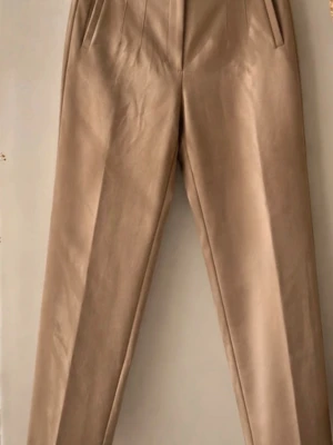 Beige kostymbyxa med pressveck - Snygga beige kostymbyxor med raka ben och pressveck framtill. Byxorna har hög midja och sidofickor, vilket ger en clean och stilren look. Perfekta till en dressad outfit och passar till många olika stilar.