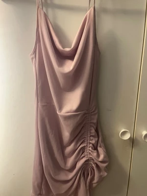 Ljuslila kort klänning med drapering - Säljer en ljuslila kort klänning med tunna axelband och snygg drapering framtill. Klänningen har en cowl-neck ringning och rynkade detaljer på sidan som ger en cool och trendig look. Perfekt för dig som vill sticka ut lite extra.