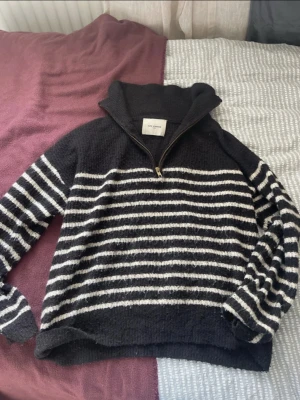 Svart stickad tröja med vita ränder - Mysig svart stickad tröja med vita horisontella ränder och halv dragkedja vid halsen. Tröjan har hög krage och långa ärmar, perfekt för kyliga dagar. Materialet ser ut att vara mjukt och fluffigt, vilket ger en avslappnad och trendig look.