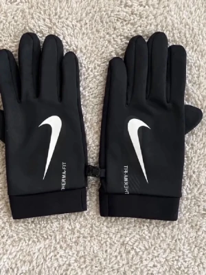 Svarta Nike Therma-Fit handskar - Snygga svarta handskar från Nike med Therma-Fit-tryck och den klassiska vita swoosh-loggan på ovansidan. Perfekta för kyliga dagar och har en sportig look. Handskarna har smidig passform och är lätta att bära med sig.