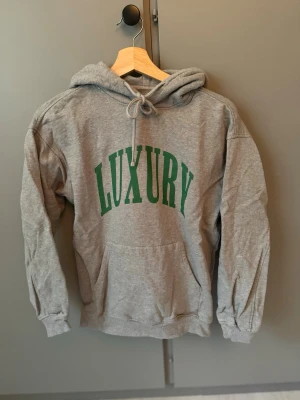 Grå hoodie med grön text LUXURY - Säljer en grå hoodie med huva och dragsko, samt stor ficka framtill. På bröstet står det 'LUXURY' i grönt tryck. Hoodien har ribbade muddar vid ärmar och nederkant. Perfekt för en chill och avslappnad stil.