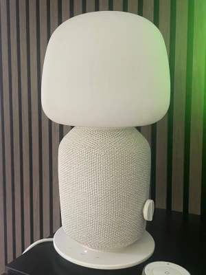 IKEA SYMFONISK Bordslampa med WiFi-högtalare och fjärrkontroll - IKEA SYMFONISK bordslampa med inbyggd WiFi-högtalare, modell E1776. Lampan är designad i samarbete med Sonos och erbjuder både belysning och ljud. Den har en vit textilklädd bas och en vit opalglas-lampskärm. En fjärrkontroll medföljer för enkel styrning av lampans funktioner. Lampan ansluts via en strömkabel och har även en Ethernet-port.