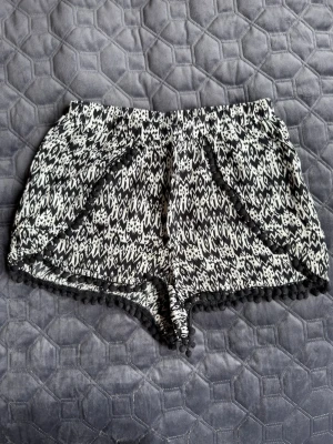 Svarta och vita mönstrade shorts - Säljer ett par svarta och vita shorts med grafiskt mönster och elastisk midja. Shortsen har dekorativa bollar längs kanten och är perfekta för varma dagar. Lätta och luftiga, passar till många olika stilar.