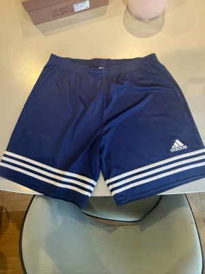 Blå Adidas shorts  - Snygga blå shorts från Adidas med klassiska vita ränder längst ner på benen och Adidas-logga på vänster ben. Shortsen har elastisk midja och är tillverkade i ett lätt och bekvämt material, perfekta för sport eller chill.