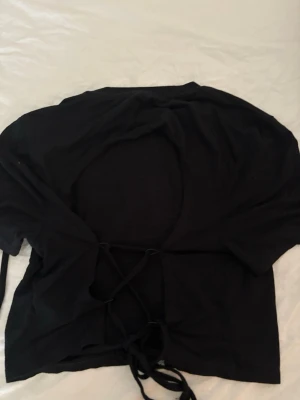 Svart bolerotopp med snörning - Trendig svart bolerotopp med öppen rygg och snörning baktill. Toppen har korta ärmar och croppad passform, perfekt för att lyfta vilken outfit som helst. Materialet känns mjukt och stretchigt, vilket gör den bekväm att bära. Öppen vid ryggen