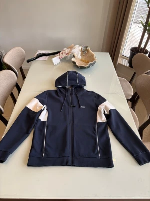Mörkblå hoodie med vita detaljer - Snygg mörkblå hoodie med vita paneler på ärmarna och vita linjer på huvan. Dragkedja framtill och två fickor. Perfekt för en avslappnad och sportig stil.