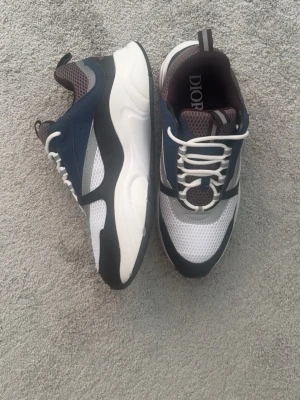 Dior vita och blå sneakers - Snygga sneakers från Dior i vitt, blått och svart med meshdetaljer och reflekterande partier. Skorna har en chunky sula och klassisk snörning. Perfekta för dig som vill ha en modern och sportig look med exklusiv känsla.