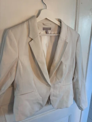 Vit kavaj från H&M - Stilren krämvit kavaj från H&M med klassisk passform och spetsiga slag. Kavajen har två fickor framtill och är helfodrad för extra komfort. Perfekt för att lyfta vilken outfit som helst med en fräsch och modern look.