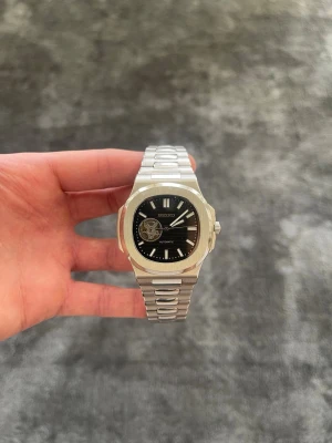 Seiko Mod Nautilus – Silver Open Heart (Svart) - ✨ Specifikationer Modell: Seiko Mod Nautilus Open Heart 🖤 Urverk: Automatiskt Seiko-urverk Gångreserv: 41 timmar ⏳ Boett: Rostfritt stål Storlek: 40 mm 📏 Glas: Reptåligt safirglas 💎 Urtavla: Svart tavla med open-heart-fönster 🫀