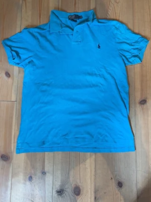 Blå pikétröja från Ralph Lauren - Säljer en blå pikétröja från Ralph Lauren i slim fit-modell. Tröjan har korta ärmar, klassisk krage och knappslå framtill. Den är tillverkad i bomull och har den ikoniska lilla loggan broderad på bröstet.