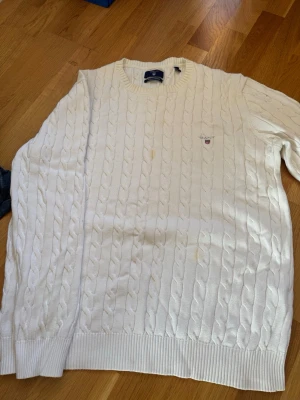 Vit kabelstickad tröja 2XL GANT  - Den har en fläck men den går att tvätta bort. Kabelstickad vit tröja med rund hals och ribbade muddar. Tröjan har ett diskret broderat märke på bröstet och är i en klassisk, oversized modell. Perfekt för dig som gillar en clean och stilren look.