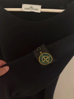 Svart stickad tröja från Stone Island - Snygg svart stickad tröja från Stone Island med klassisk rund halsringning och ikonisk logotyp på vänster ärm. Tröjan har en stilren och enkel design med långa ärmar och ribbade muddar. Perfekt för dig som gillar streetwear och exklusiva detaljer.