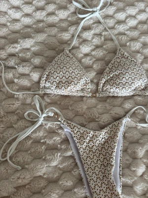 Vit och beige bikini med knyt - Säljer en snygg trekantsbikini i vitt och beige med små blommiga detaljer. Både överdel och underdel har knyt i sidorna och nacken.