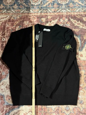 Svart stickad tröja från Stone Island - Svart stickad tröja från Stone Island med klassisk rund hals och ribbade muddar. Tröjan har den ikoniska Stone Island-loggan på vänster ärm och är tillverkad i ett mjukt, bekvämt material. Perfekt för dig som gillar stilrena och tidlösa plagg.
