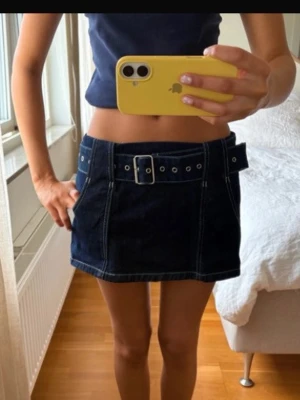 Mörkblå jeanskjol med bälte - Snygg mörkblå jeanskjol med markerade sömmar och ett brett bälte med metallspänne. Kjolen har fickor fram och en kort, A-linjeformad passform som ger en trendig look. Perfekt att matcha med en enkel topp för en cool outfit.