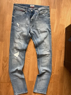 Ljusblå Tommy jeans i slim tapered passform.  - Säljer ett par ljusblå jeans med slitningar och distressed detaljer framtill. Jeansen har fem fickor, knappgylf och smal passform.