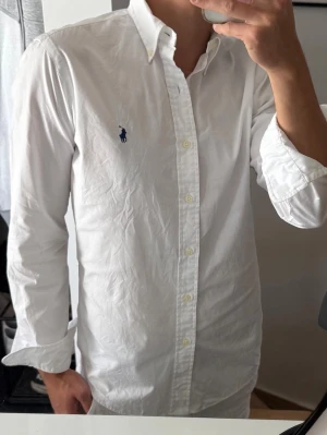 Vit skjorta från Ralph Lauren - Klassisk vit skjorta från Ralph Lauren med blå broderad logga på bröstet. Skjortan har lång ärm, knappar framtill och en tidlös passform. Tillverkad i bomull för en skön känsla och stilren look.