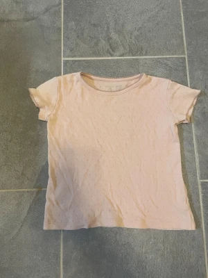 Ljusrosa t-shirt med hålmönster - Söt ljusrosa t-shirt för barn med korta ärmar och rund hals. T-shirten har ett diskret hålmönster över hela tyget som ger en extra detalj. Perfekt till vardags och enkel att matcha med olika stilar.