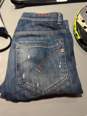 Blå jeans med slitningar - Säljer ett par klassiska blå jeans med snygga slitningar och kontrastsömmar. Jeansen har fem fickor och en cool detalj på bakfickan med orange söm. Perfekta för en avslappnad och trendig look.
