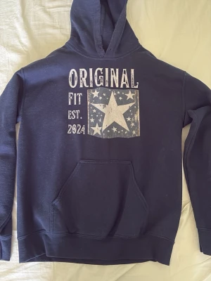 Mörkblå hoodie med stjärntryck - Snygg mörkblå hoodie med vit text och stort stjärntryck på bröstet. Texten säger 'ORIGINAL FIT EST. 2024'. Hoodien har klassisk känguruficka och huva. Perfekt för en avslappnad streetwear-look.