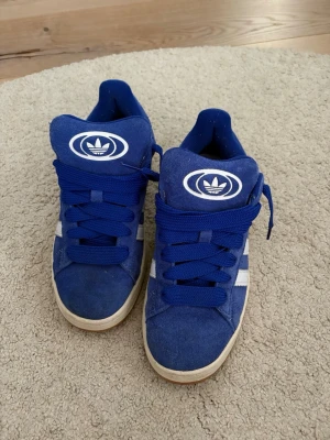 Adidas campus - Snygga blå sneakers från Adidas med klassiska vita ränder på sidorna och stor Adidas-logga på plösen. Skorna har breda blå skosnören och en vit sula. Ovandelen är i mockaliknande material och modellen är låg.