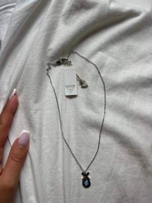 Guess silverhalsband - Snyggt halsband från Guess i silverfärgad metall med en rund blå sten i mitten av ett cirkelformat hänge och ett litet X ovanför. Kedjan är tunn och har en liten Guess-logga vid låset. Perfekt för dig som gillar stilrena accessoarer.