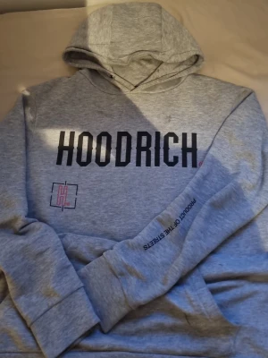 HOODRICH hoodie - Snygg grå hoodie från HOODRICH med stor svart logga på bröstet och textdetaljer på ärmen. Klassisk huva och känguruficka framtill. Tillverkad i mjukt sweatshirtmaterial som är perfekt för en avslappnad streetwear-look.OBS! har en liten fläck (syns knappt)