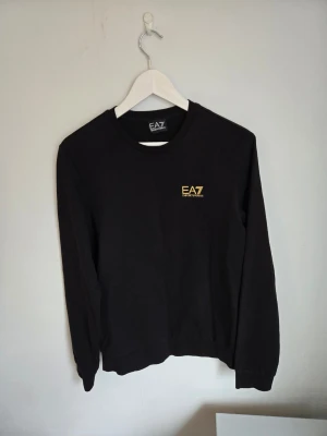 Svart EA7 sweatshirt med guld logga - Snygg svart sweatshirt från EA7 med gul logga på bröstet och guld detalj på ryggen. Tröjan har rund halsringning, långa ärmar och ribbade muddar. Perfekt för en clean och sportig look.