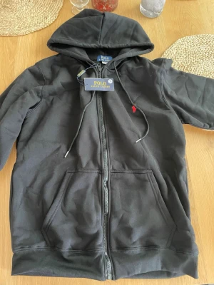 Polo Ralph Lauren hoodie - Svart hoodie med dragkedja från Polo Ralph Lauren. Klassisk design med huva, dragsnören och kängurufickor. Liten röd logga broderad på bröstet. Perfekt för en avslappnad och stilren look. Riktigt sköna! Storlek M. Kom privat vid minsta fundering!