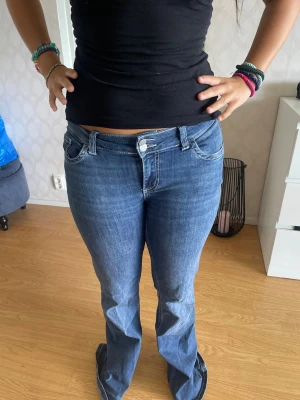 Blå bootcut jeans med kontrastsömmar - Säljer ett par blå bootcut jeans med vita kontrastsömmar och fickor bak med broderade detaljer. Jeansen har låg midja och klassisk femficksdesign. Perfekta för dig som gillar en avslappnad men ändå snygg stil.