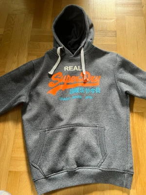 Grå hoodie från Superdry med tryck - Säljer en grå hoodie från Superdry med vita snören. Framsidan har ett färgglatt tryck i orange och turkos med Superdry-logga och japansk text. Insidan är mjuk och fleecefodrad, perfekt för chill dagar. Trycket är lite slitet men annars är allt väldigt bra 
