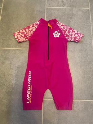 Rosa UV-baddräkt med blommönster - Säljer en rosa UV-baddräkt med korta ben och ärmar. Ärmarna har vitt blommönster och framtill finns en vit hibiskusblomma. Dragkedja framtill och texten 'LIFEGUARD Smiling Shark' på benet. Perfekt för stranden eller poolen.