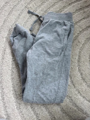 Grå mjukisbyxor från Kappahl - Mysiga grå mjukisbyxor från Kappahl med resår och dragsko i midjan samt ribbade muddar vid bensluten. Tillverkade i mjukt och skönt material, perfekta för chill dagar hemma eller till träningen.