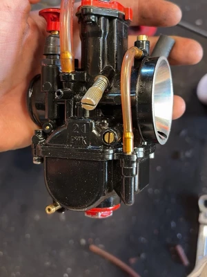 Svart 21mm PWK förgasare - Säljer en svart 21mm PWK förgasare med röda och mässingsfärgade detaljer samt genomskinliga bränsleslangar. Passar till moped eller motorcykel och har en rund insugskoppling i metall. Perfekt för dig som vill trimma eller byta ut din nuvarande förgasare.