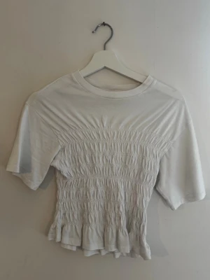 Vit smockad topp med volangkant - Söt vit topp med smockad midja och volangkant nertill. T-shirten har korta ärmar och rund halsringning. Perfekt för en trendig och avslappnad look. Pris går att diskutera💕💕