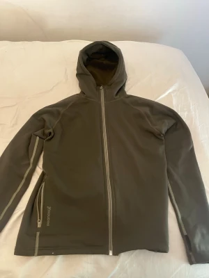 Houdini hoodie - Säljer en olivgrön hoodie från Houdini med hel dragkedja framtill och huva. Hoodien har långa ärmar och diskreta fickor med dragkedja på sidan. Tillverkad i mjukt och stretchigt material, perfekt för chill eller äventyr.