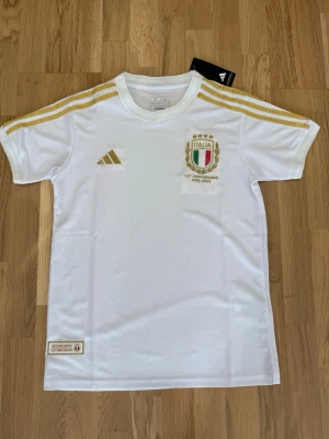 Italien tröja - Säljer en vit Italien jubileumströja från Adidas med guldfärgade detaljer och tre ränder på axlarna. Tröjan har Italiens klubbmärke på bröstet och är tillverkad i ett lätt, ventilerande material. Perfekt för fotboll eller träning.