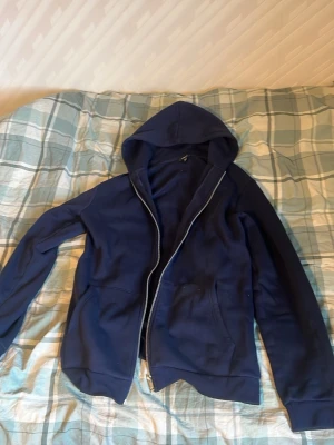 Mörkblå zip hoodie med huva - Säljer en mörkblå hoodie med dragkedja och huva. Tröjan har två fickor framtill och ribbade muddar vid ärmslut och nederkant. Perfekt för en avslappnad stil och enkel att matcha med jeans eller joggers.
