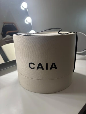 Beige rund necessär/box från CAIA - Säljer en beige rund necessär/box från CAIA med svart handtag och tydlig logga framtill. Den har en hård struktur och är perfekt för att förvara smink eller accessoarer. Stilren design som sticker ut på sminkbordet.