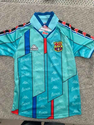 Turkos FC Barcelona tröja från Kappa - Säljer en turkos FC Barcelona matchtröja från Kappa med klubbmärke på bröstet och röda samt blå detaljer. Tröjan har korta ärmar, klassisk krage och tryckta loggor längs axlarna. Perfekt för dig som älskar fotboll och vill sticka ut på planen.