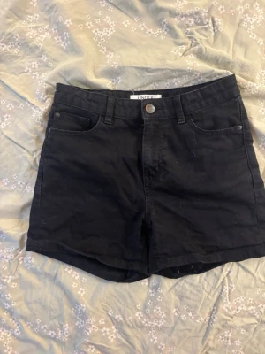 Svarta höga jeanshorts - Säljer ett par svarta, högmidjade shorts i mjuk jeansmaterial. De har klassisk femficksdesign, knapp och dragkedja framtill samt en tight passform som framhäver midjan. Man kan välja mellan att ha de i sin orginella passform eller vika upp benen så det blir minishorts. Perfekta för varma sommardagar och enkla att matcha med olika toppar.